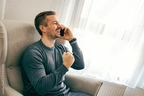A young man is receiving great news on the phone while sitting at his comfortable home.