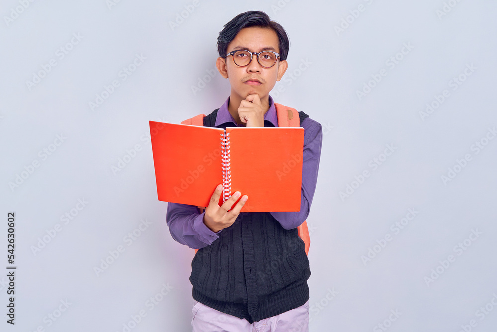 Concentrated young Asian students with backpacks holding books, thinking about question and answer isolated on gray background. Education in high school university college concept Concentrated young Asian students with backpacks holding books, thinking about question and answer isolated on gray background. Education in high school university college concept