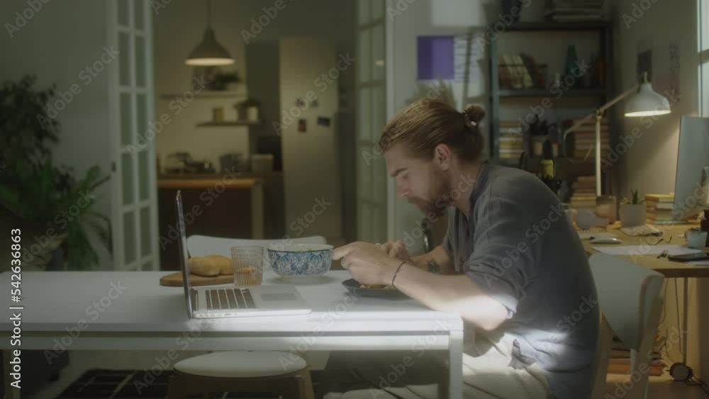 Side view of young bearded man eating breakfast and watching movie on laptop in morning at home Side view of young bearded man eating breakfast and watching movie on laptop in morning at home