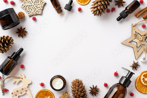 Winter skin care concept. Top view photo of glass cosmetic bottles wooden ornaments pine cones mistletoe berries anise and dried orange slices on isolated white background with copyspace in the middle