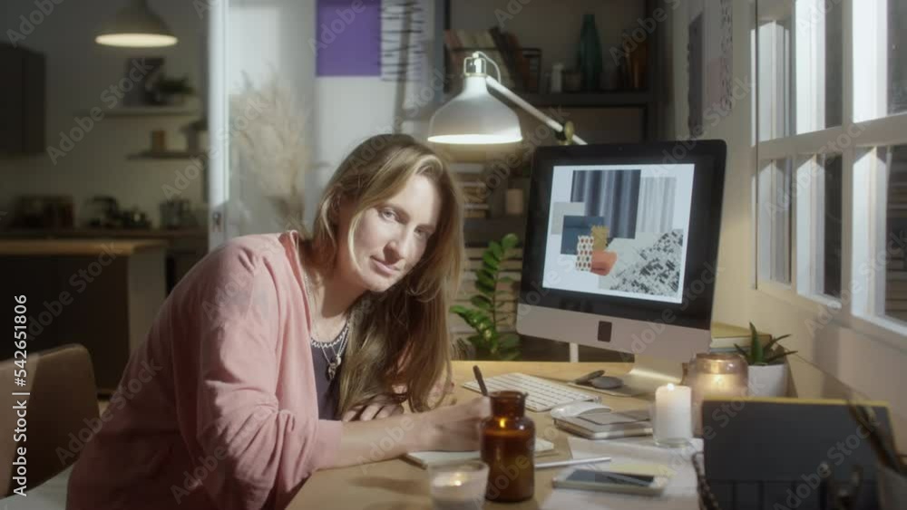 Portrait of young female designer taking notes at desk with burning candles and then posing for camera while working in cozy atmosphere at home Portrait of young female designer taking notes at desk with burning candles and then posing for camera while working in cozy atmosphere at home