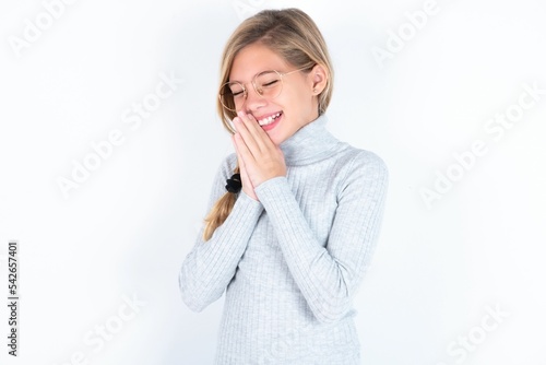 Overjoyed beautiful caucasian teen girl wearing gray turtleneck sweater over white wall laughs joyfully and keeps palms pressed together hears something funny