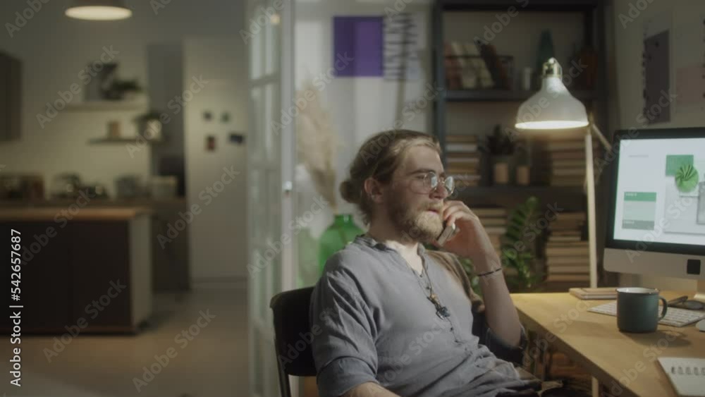 Medium shot of male freelancer sitting at home workplace in evening and making phone call Medium shot of male freelancer sitting at home workplace in evening and making phone call