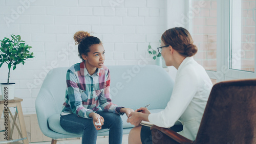 Emotional African American lady is talking to psychologist sitting on couch, talking and gesturing during professional consultation while specialist is listening holding paper and pen.
