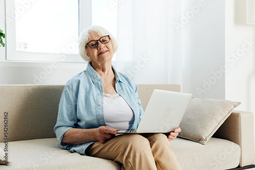 a happy, joyful, aged woman is sitting in a beautiful bright apartment with a laptop on a cozy beige sofa and looks with a pleasant smile on her face © Tatiana