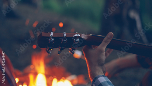 Close-up shot of male tourist's hand playing the guitar during romantic evening at campsite with fire burning in background. Musical instruments, nature and people concept.