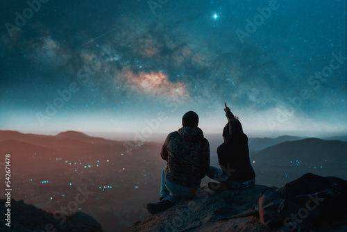 silhouette of a couple sitting on top of a hill looking at the stars over the city  back view