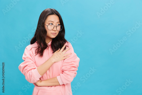 Confused sad cute Asian young lady in pink hoodie sweatshirt wear round glasses looks aside posing isolated on over blue studio background. The best offer for ad. Eyewear for vision correction concept