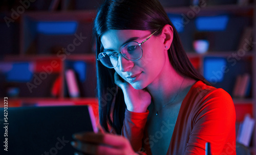 Focused on the work. Beautiful woman in glasses and red wear is sitting by the laptop in dark room with neon lighting