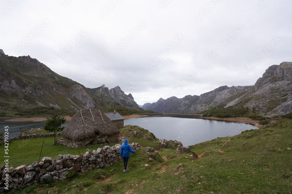 Woman with a raincoat in front of a cabin with a vegetable roof walks towards the lake in a beautiful natural setting in Asturias. Somiedo Natural Park and Biosphere Reserve.