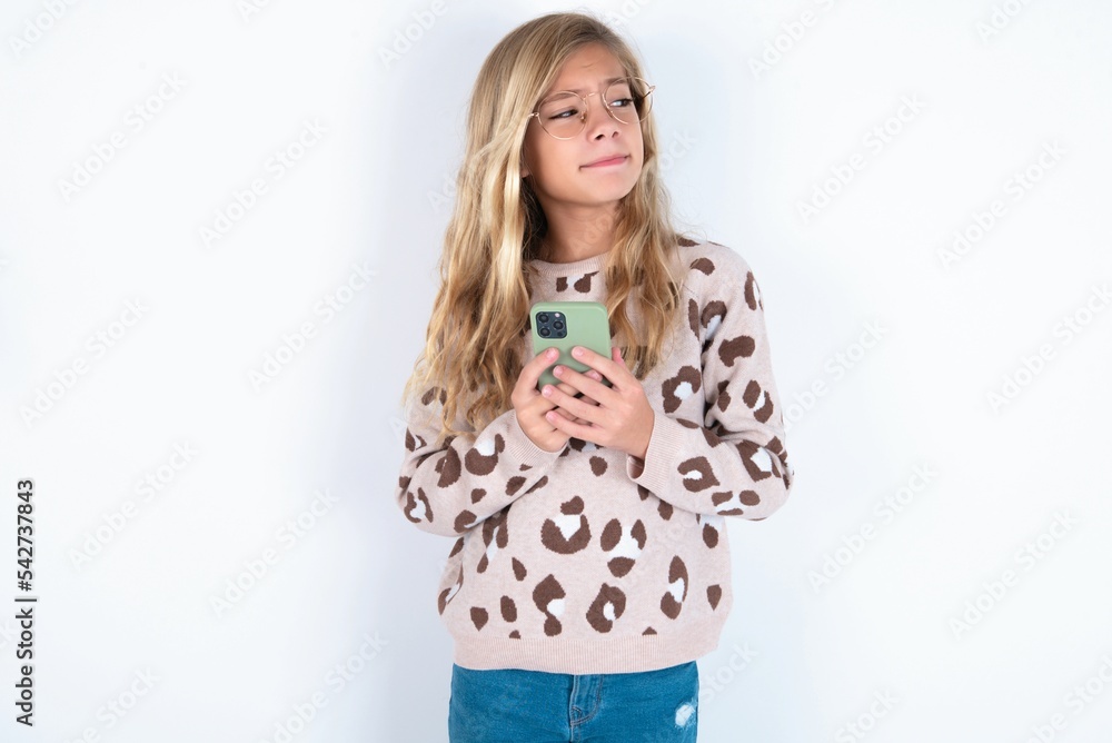 Portrait of beautiful caucasian teen girl wearing animal print sweater over white wall with dreamy look, thinking while holding smartphone. Tries to write up a message. Portrait of beautiful caucasian teen girl wearing animal print sweater over white wall with dreamy look, thinking while holding smartphone. Tries to write up a message.