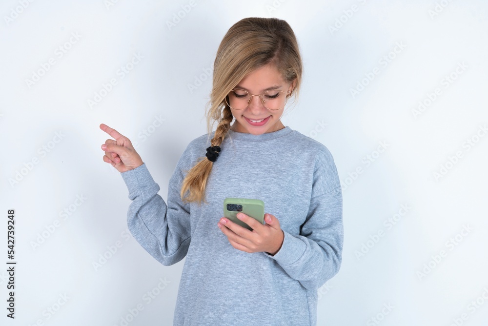Smiling beautiful caucasian teen girl wearing gray sweater over white wall pointing finger at blank space holding phone in one hand Smiling beautiful caucasian teen girl wearing gray sweater over white wall pointing finger at blank space holding phone in one hand