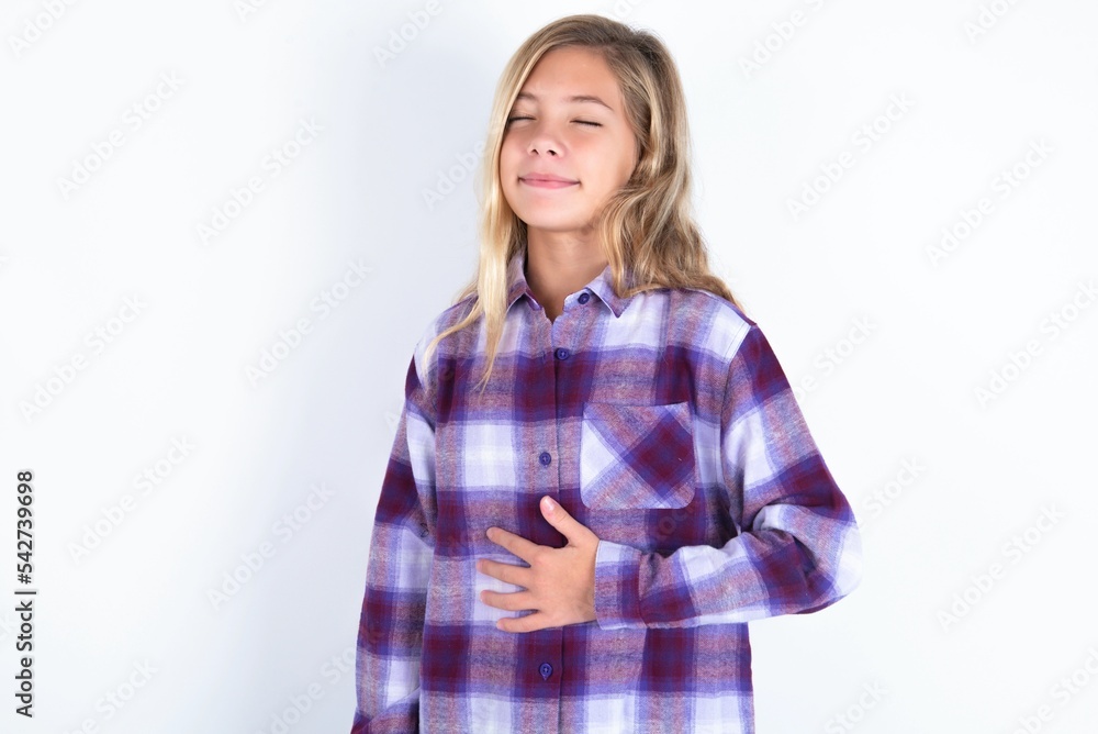 beautiful caucasian teen girl wearing plaid purple shirt over white wall touches tummy, smiles gently, eating and satisfaction concept. beautiful caucasian teen girl wearing plaid purple shirt over white wall touches tummy, smiles gently, eating and satisfaction concept.