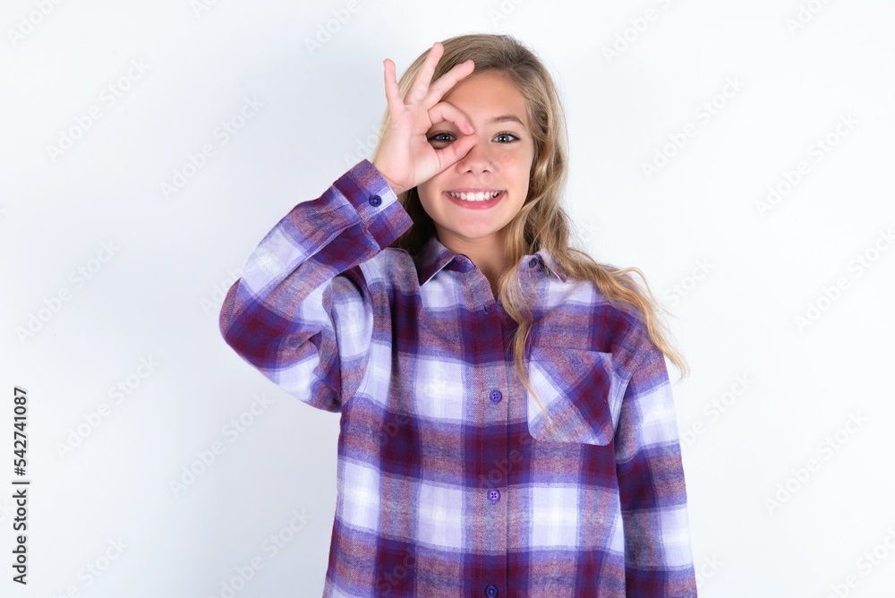 beautiful caucasian teen girl wearing plaid purple shirt over white wall doing ok gesture with hand smiling, eye looking through fingers with happy face. beautiful caucasian teen girl wearing plaid purple shirt over white wall doing ok gesture with hand smiling, eye looking through fingers with happy face.