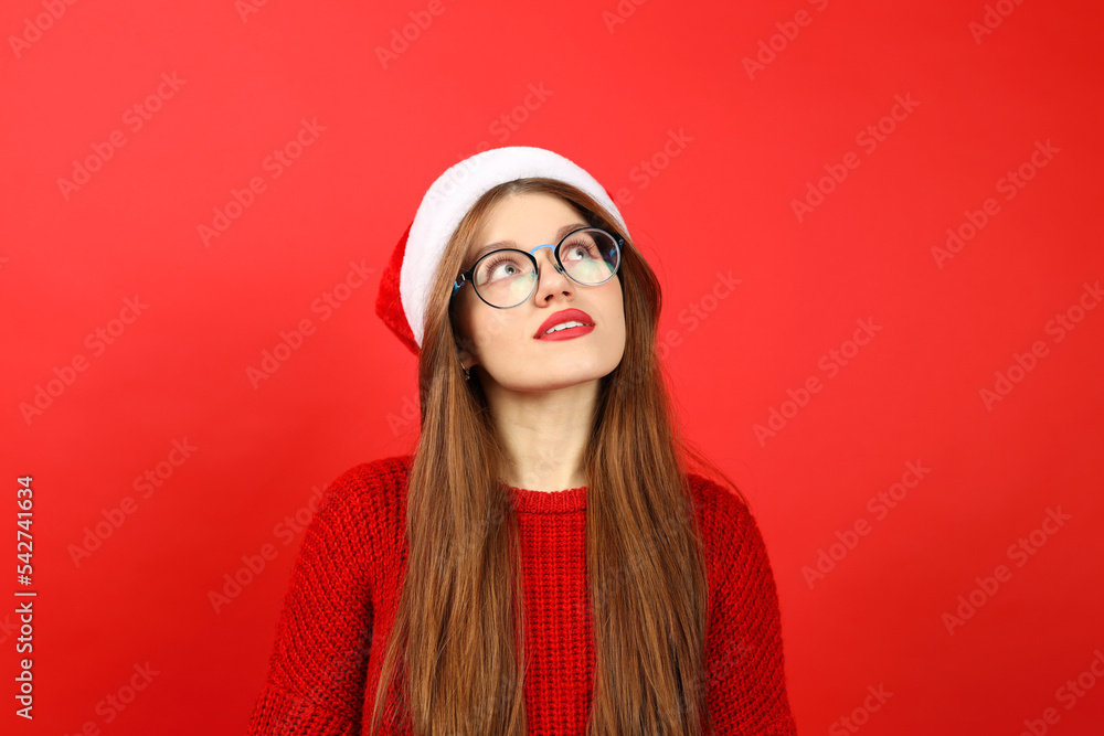 Portrait of an attractive girl in a santa hat and a Christmas sweater thinks about choosing a New Year's gift. Woman dreaming on a red background. Portrait of an attractive girl in a santa hat and a Christmas sweater thinks about choosing a New Year's gift. Woman dreaming on a red background.