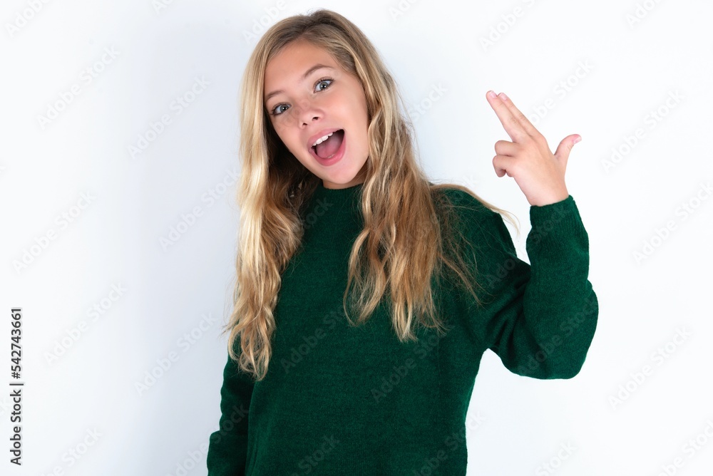 beautiful caucasian teen girl wearing green knitted sweater over white wall foolishness around shoots in temple with fingers makes suicide gesture. Funny model makes finger gun pistol beautiful caucasian teen girl wearing green knitted sweater over white wall foolishness around shoots in temple with fingers makes suicide gesture. Funny model makes finger gun pistol
