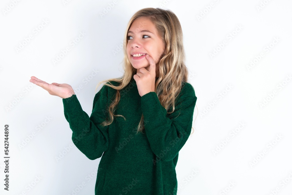 Obraz premium Positive beautiful caucasian teen girl wearing green knitted sweater over white wall advert promo touch finger teeth