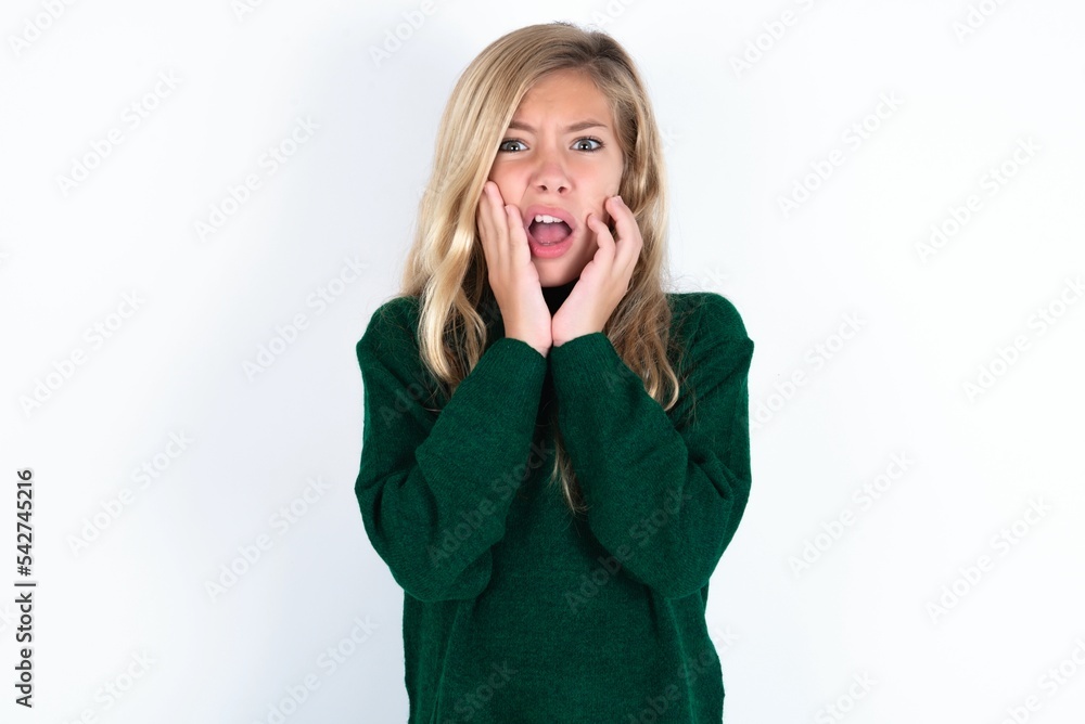 Upset beautiful caucasian teen girl wearing green knitted sweater over white wall touching face with two hands Upset beautiful caucasian teen girl wearing green knitted sweater over white wall touching face with two hands