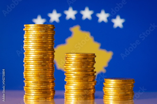 Problems in economy of Kosovo concept. Descending graph made of gold coins stacks with flag of Kosovo on the background