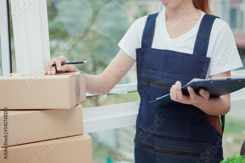 Asian woman business owner with many parcel boxes on table Happy with online sales, use smartphone, calculator and paper to work. Receive orders from customers, take notes, and arrange shipments.