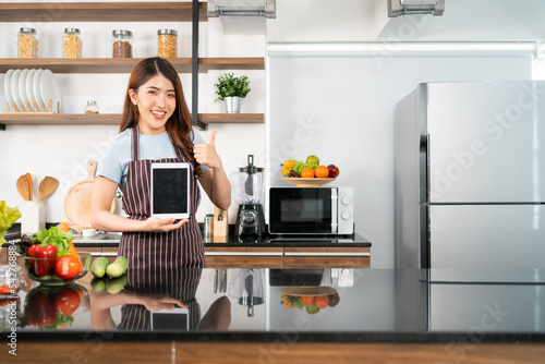 A happy Asian woman wearing an apron shows a digital tablet in her home kitchen. Prepares vegetables for healthy food by searching recipes online. Modern cooking with technology.