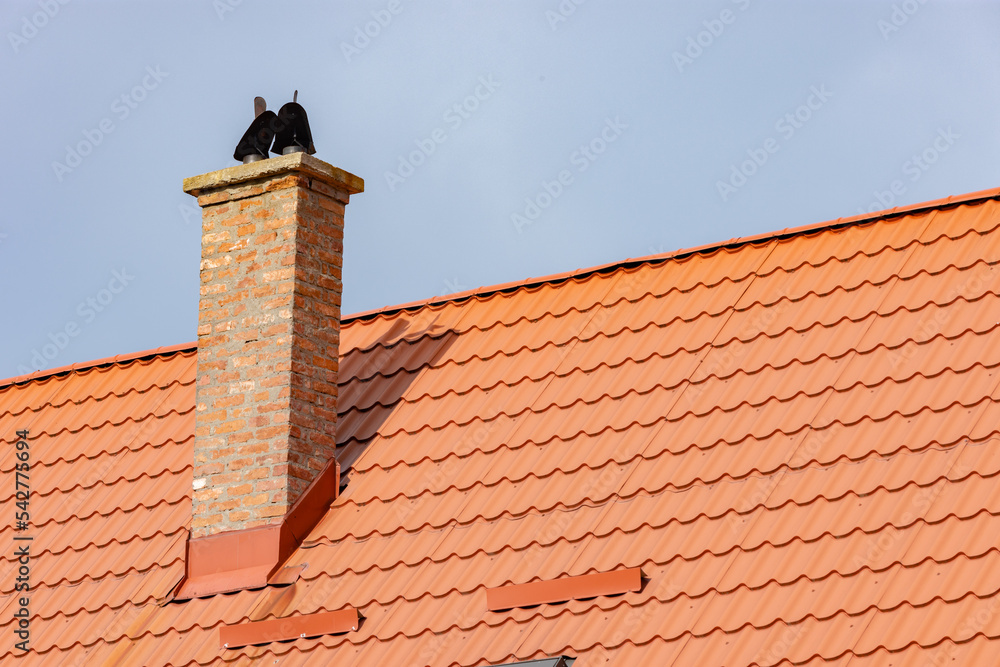 Restored roof with orange tiles and a chimney made of bricks and cement with two open cowls for rain protection against a clear blue sky