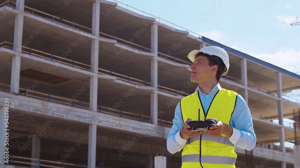 Drone operator holding remote controller. Professional builder in helmet and vest standing in front of construction site. Office building and crane background. Business, real estate and investment.