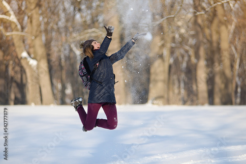 Happy young woman jumping and throwing up snow in snowy winter park. Sunny weather. Winter season.