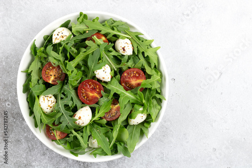 Green salad with arugula, cherry tomatoes, mozzarella cheese, olive oil and spices in a white plate on a gray background. Top view.