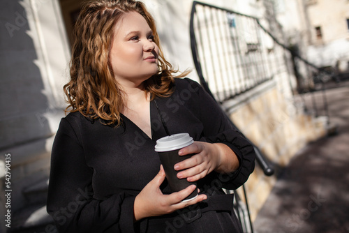 Overweight woman with cup of coffee or tea walking outdoor in the street. Plus size girl wearing black dress, is holding the packege with purchases photo