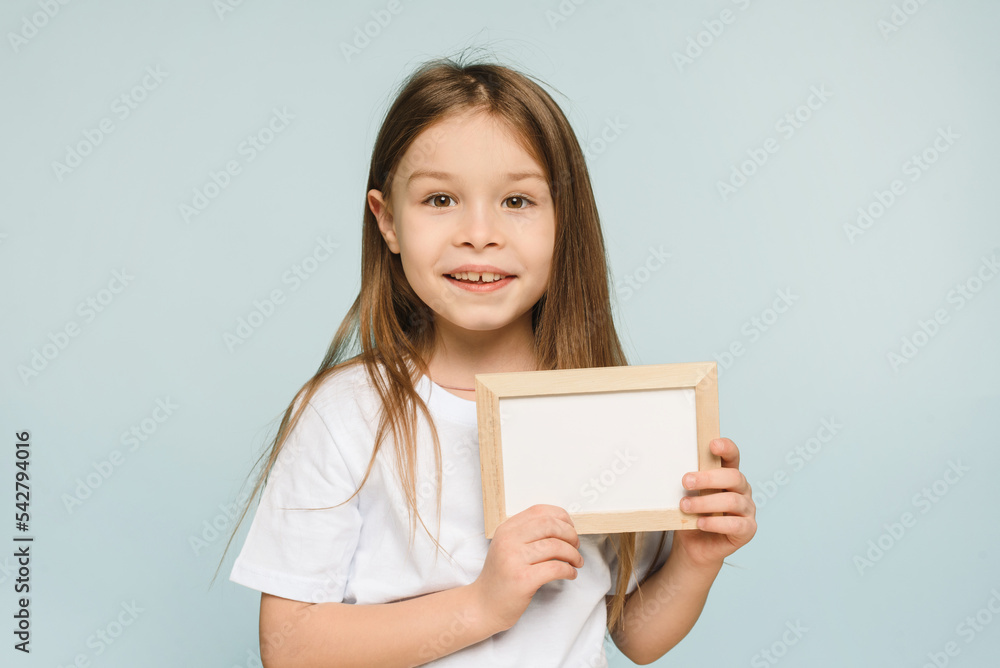 Close up little girl holding white blank frame posing isolated on pastel blue background in studio. Copy space mockup Close up little girl holding white blank frame posing isolated on pastel blue background in studio. Copy space mockup