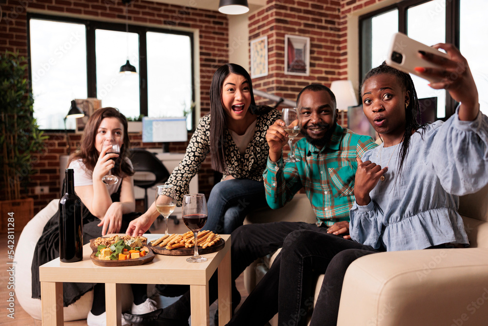 Multi ethnic young adults group posing for mobile selfie at apartment living room, smiling, laughing, tasting snacks drinking wine. African american woman photographing friends.