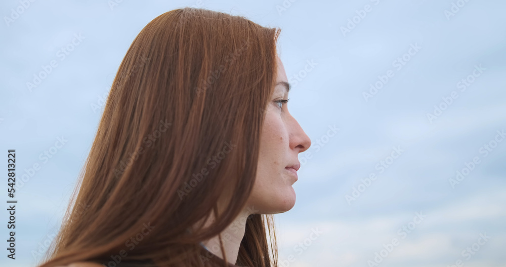 Red-haired young woman looks thoughtfully into the distance against a cloudy sky. Concept of appeasement, tranquility, peace, anxiety. Red-haired young woman looks thoughtfully into the distance against a cloudy sky. Concept of appeasement, tranquility, peace, anxiety.