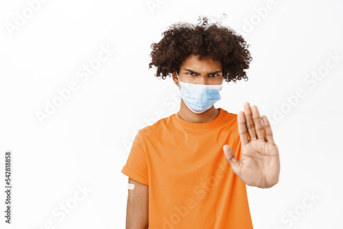 Man showing stop sign, wearing medical face mask, showing thumbs down, he has patch on arm after covid-19 vaccination, dislike smth, standing over white background © Cookie Studio