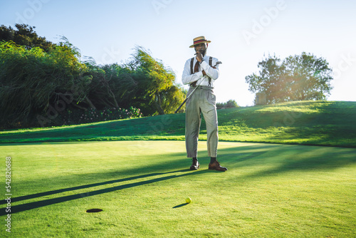 View of a fashionable handsome bearded black senior with a sigar, in straw hat and tailored outfit standing on a green golf field with a club in his hand; a yellow golf ball and a hole in front of him photo