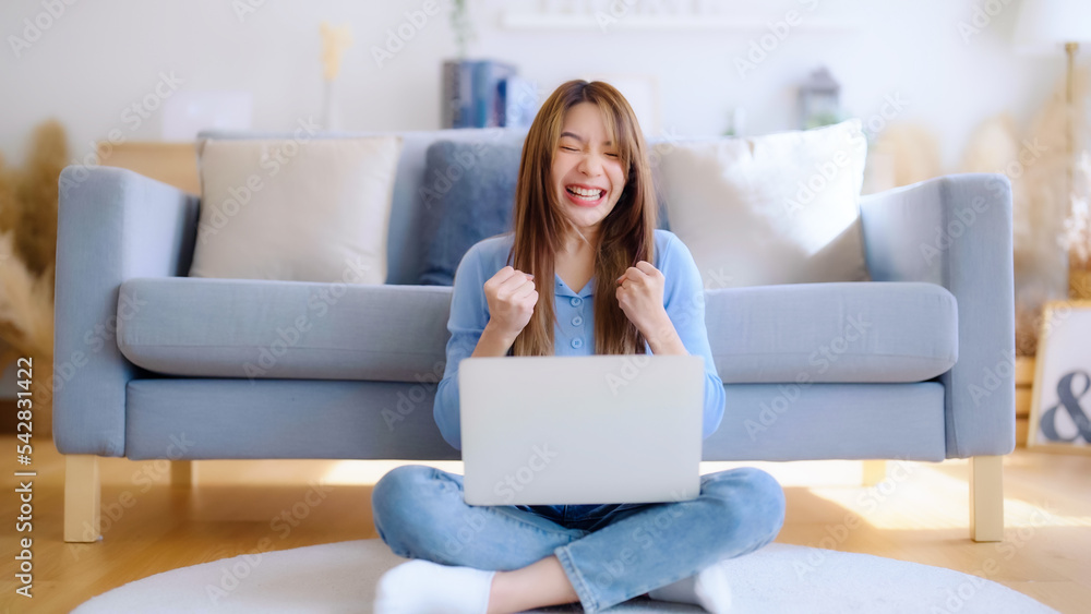 Young asian woman in good spirits working on laptop at home while sitting on the floor close to the couch. Excited female winner celebrating success while glancing at notebook Young asian woman in good spirits working on laptop at home while sitting on the floor close to the couch. Excited female winner celebrating success while glancing at notebook