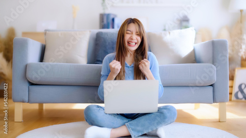 Young asian woman in good spirits working on laptop at home while sitting on the floor close to the couch. Excited female winner celebrating success while glancing at notebook