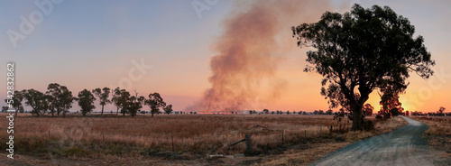 Panoramic image of a plume of balck smoke rising at sunset from a line of wild fire in the distant farmland, a gravel road and native trees in the foreground, rural Victoria, Australia