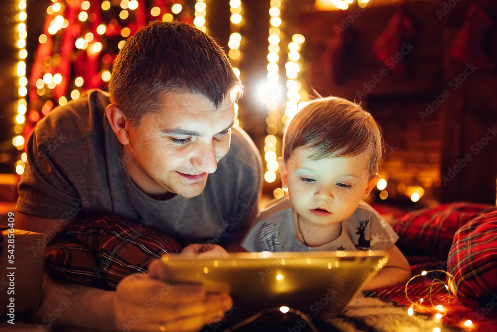 Dad and little son using tablet pc at home near Christmas tree in cozy living room on Christmas eve