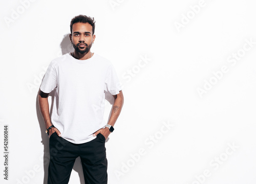 Handsome hipster model. Sexy unshaven man dressed in summer t-shirt and jeans clothes. Fashion male posing near white wall in studio at sunny day. Isolated