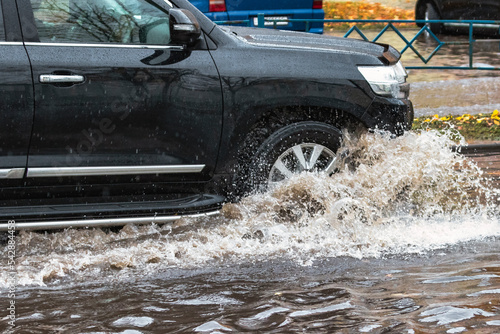 The car is driving through a puddle in heavy rain. Splashes of water from under the wheels of a car. Flooding and high water in the city.