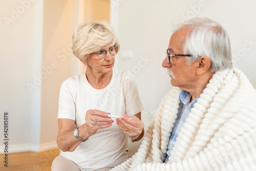 Woman checking fever temperature of senior man sitting on bed. Mature husband resting at home feeling symptoms of the flu while wife checking fever.