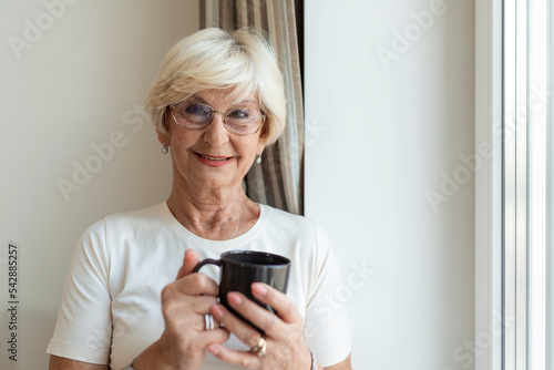 Happy senior woman day dreaming while looking through the window and enjoying in cup of coffee. Positive mature woman in white cotton blouse drinking mug of coffee and looking through window