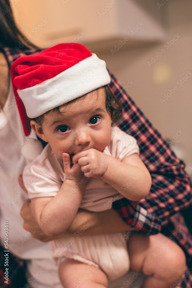 Mother is holding a baby in a Santa hat at home,close-up.Preparing for Christmas.Upcoming winter holidays and celebrations. Mother is holding a baby in a Santa hat at home,close-up.Preparing for Christmas.Upcoming winter holidays and celebrations.