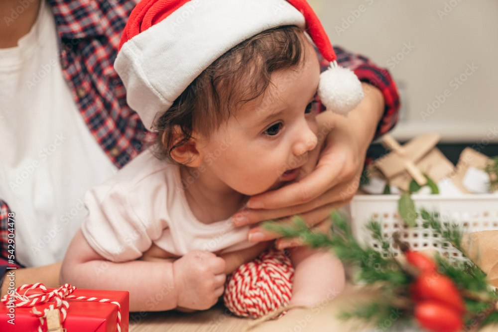 Mother is holding a baby in a Santa hat at home,close-up.Preparing for Christmas.Upcoming winter holidays and celebrations. Mother is holding a baby in a Santa hat at home,close-up.Preparing for Christmas.Upcoming winter holidays and celebrations.