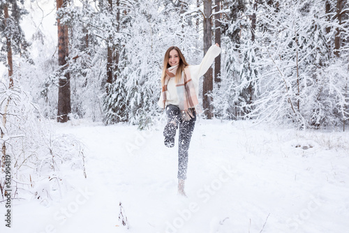 Girl in warm scarf and white sweater having fun kicking up snow with her leg outdoors in winter snowy forest 