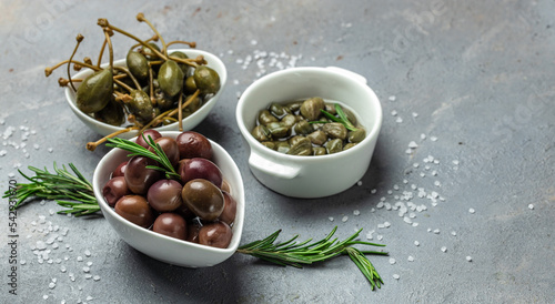 Foto Marinated Green and Kalamata Olives and Capers with fresh rosemary in a small bowl, on light wooden background