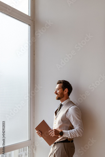 Side view of positive businessman in formal wear holding paper folder near window in office 