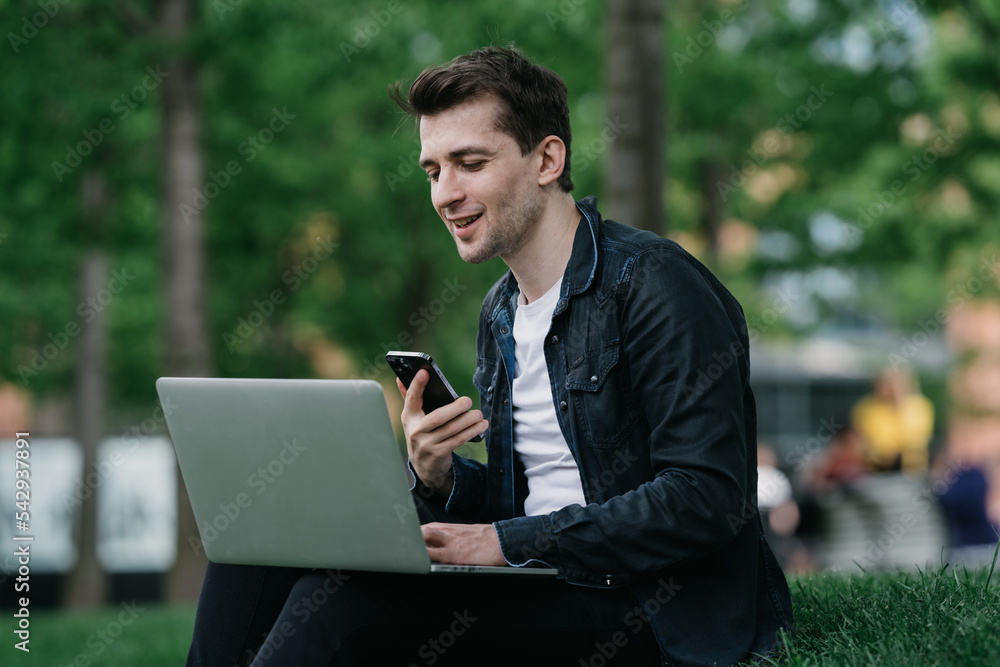 Handsome brunette cheerful male sitting on grass at park with laptop making video call holds phone smiling having break remote working. Cheerful man student talking with friend via internet.