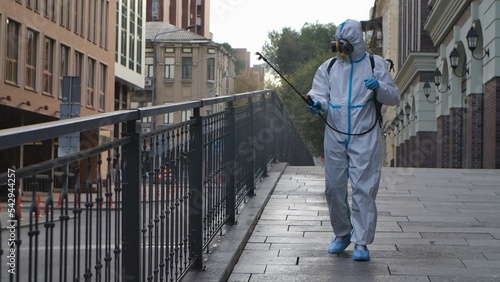 Medical worker in white protective suit and respirator disinfects railing on city street. Virologist treating objects with an antiseptic using an alcohol disinfectant and sprinkler.